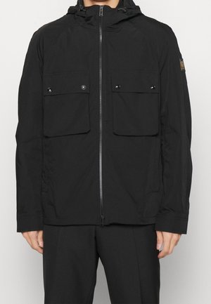 Homme portant une veste noire à capuche zippée avec deux poches poitrine à boutons et un pantalon noir, debout devant un fond uni.