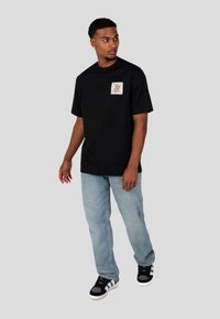 Czarny bawełniany T-shirt z kwadratową kieszonką graficzną, w połączeniu z jasnoniebieskimi dżinsami i czarnymi sneakersami adidas.