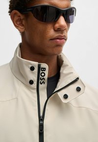 Giacca impermeabile beige con colletto alto, zip nera e bottoni. Presenta il logo "BOSS" sul colletto e una finitura liscia e testurizzata.