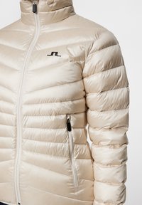 Jasnobeżowa kurtka puchowa z wysokim kołnierzem, pikowanym wzorem, zamek błyskawiczny z przodu i boczną kieszenią. Na klatce piersiowej czarna logo.
