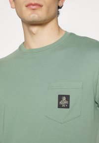 Ljusgrön bomullströja med rund hals, korta ärmar och en bröstficka med en svart etikettdesign. Slät textur.