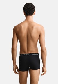 Männliches Modell von hinten gesehen, trägt schwarze GANT Boxer-Shorts vor weißem Hintergrund, steht mit entspannt hängenden Armen an der Seite.