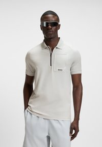 Man wearing light beige short-sleeve polo shirt with zip and "BOSS" logo pocket, white shorts, and reflective wrap sunglasses, standing with hands in pockets.