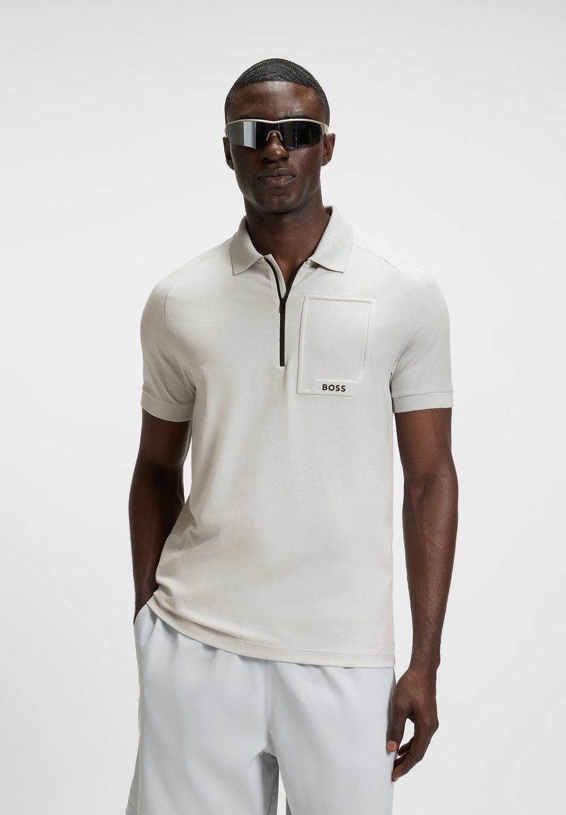 Man wearing light beige short-sleeve polo shirt with zip and "BOSS" logo pocket, white shorts, and reflective wrap sunglasses, standing with hands in pockets.