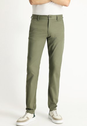 Pantaloni slim-fit verde oliva realizzati in tessuto liscio, con chiusura a bottone, tasche laterali e abbinati a sneakers bianche e grigie.