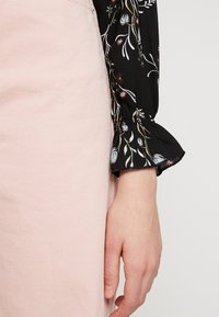 Fluffig svart blus med blommönster och samlat detalj vid handleden, ihop med en ljusrosa denimkjol. Fokusera på textilmönster och färgkontrast.