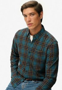 Camisa de cuadros en teal, amarillo y marino, hecha de algodón. Presenta un cuello abotonado, mangas largas y un bolsillo en el pecho. Se lleva con jeans azules.