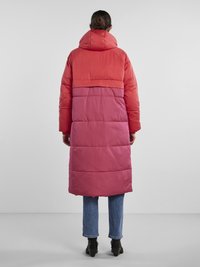 Casaco acolchoado com capuz, apresentando um design em bloco de cor em vermelho e rosa, e textura acolchoada. Usado sobre jeans azuis e botas pretas.