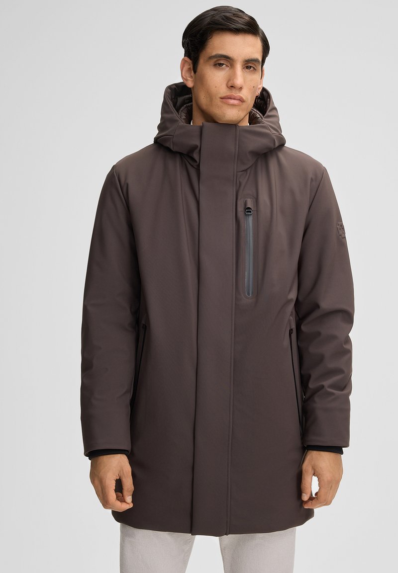 Mann trägt einen dunkelbraunen Wintermantel mit Kapuze, der einen Reißverschluss-Brusttasche und Seitentaschen hat, stehend vor einem schlichten hellen Hintergrund.