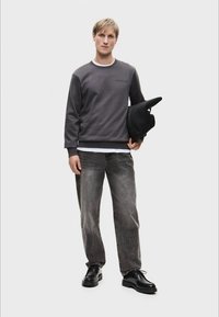 Szary sweatshirt z okrągłym dekoltem i logo, w połączeniu z jasnoszarymi dżinsami i czarnymi butami. Model trzyma czarną czapkę beanie, prosty tle.