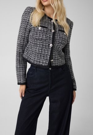 Le mannequin porte une veste en tweed noir et blanc à boutons argentés et un jean foncé à jambes larges, sur un fond gris uni.
