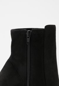 Bottine en daim noir avec fermeture éclair latérale, texture lisse et détails cousus en haut. Silhouette simple et effilée.