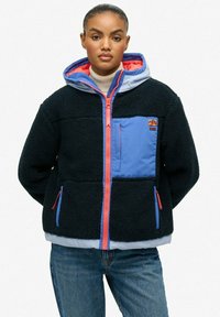 Vrouw in zwart-blauwe fleecejack met ritssluiting en koraalkleurige accenten, met capuchon, in combinatie met blauwe jeans, staand voor een witte achtergrond.