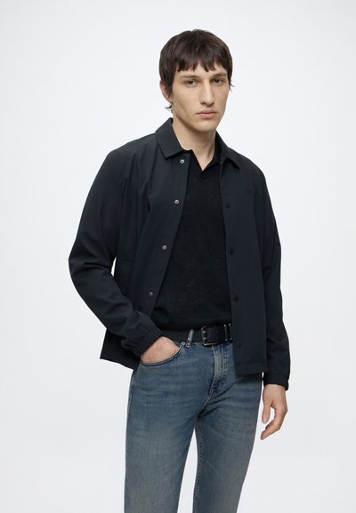 Jeune homme aux cheveux courts et foncés portant une veste noire boutonnée, une chemise noire, un jean bleu et une ceinture noire, debout devant un fond uni.