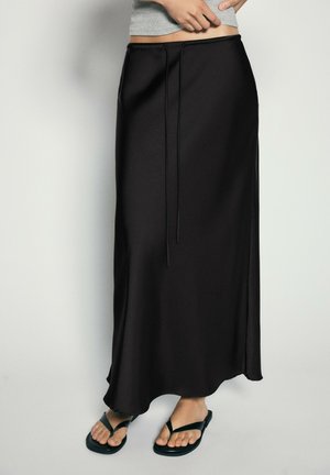 Personne portant une longue jupe noire à taille coulissante et des tongs noires, debout devant un fond clair.