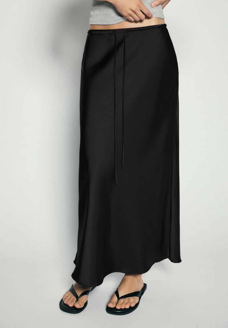 Personne portant une longue jupe noire à taille coulissante et des tongs noires, debout devant un fond clair.