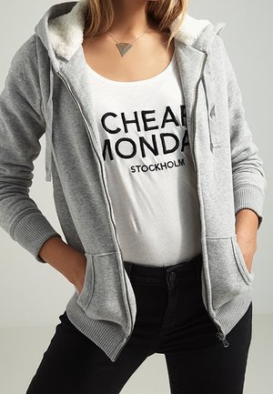 Nainen, jolla on päällään valkoinen "Cheap Monday Stockholm" t-paita, harmaa vetoketjuhuppari taskuilla, mustat housut ja kolmiomuotoinen riipuskaulakoru.