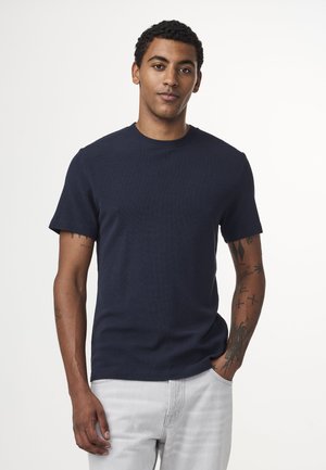 Koningblauwe T-shirt met korte mouwen, ribbelstructuur en ronde halslijn, gecombineerd met lichtgrijze jeans. Geaccentueerd door zichtbare tatoeages op de arm.