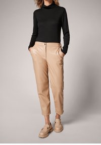 Col roulé noir ajusté, pantalon fuselé en simili cuir beige avec poches latérales. Chaussures beige à enfiler avec accessoires décoratifs. Texture lisse.