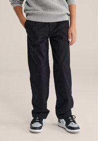 Čierne nohavice so rovnými nohavicami a hladkou textúrou, kombinované s čierno-sivými športovými teniskami. Svetre sú svetlosivé s textúrovaným pletením.