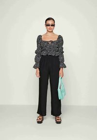Blusa preta com padrão floral e mangas bufantes, calças largas pretas e sandálias plataforma pretas. Segurando uma mala azul clara.