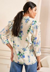 Blusa trasparente a fantasia floreale con spalle arricciate e texture a pois. Sfondo crema chiaro con fiori blu e verdi. Maniche a tre quarti.