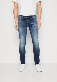 Man som bär slim-fit blåtvättade jeans, vita sneakers och en enkel vit t-shirt, stående mot en neutral bakgrund.