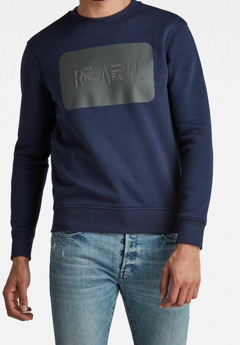 Homme portant un sweat-shirt bleu marine avec un patch rectangulaire noir marqué « R A W. » et un jean bleu clair, recadré aux épaules et aux cuisses.