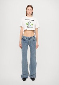 Hvit kort t-skjorte med grafisk trykk, kombinert med flagrende lys blå jeans. Modellen har på seg svarte sko. Enkel bakgrunn.