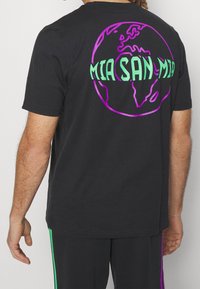 Schwarzes Baumwoll-T-Shirt mit einem bunten Globusdesign auf der Rückseite und dem Text "MIA SAN MIA" in Grün und Lila. Kurzarm, Rundhalsausschnitt.