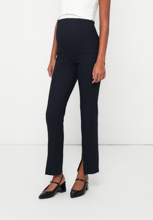 Pantaloni premaman neri a coste con vestibilità slim, dotati di slitte laterali all'orlo. Abbinati a scarpe nere con tacco a blocco.