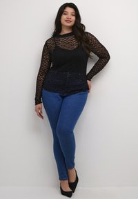 Schwarzes Spitzentop mit langen Ärmeln und geometrischen Mustern, kombiniert mit blauen Skinny-Jeans. Schwarze spitze Schuhe runden das Outfit ab.