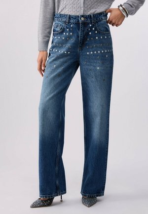 Kobieta ubrana w niebieskie dżinsy z wysokim stanem ozdobione srebrnymi ćwiekami na przedniej górnej części, w połączeniu z szarym swetrem i wzorzystymi, spiczastymi szpilkami.