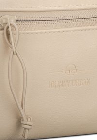 Gros plan d'une pochette à fermeture éclair en cuir beige avec logo embossé "Johnny Urban" et tirette à boucle.