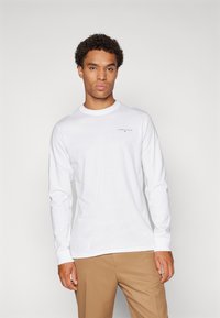 Camisola de manga longa branca em algodão com gola redonda, apresentando um pequeno logótipo preto no peito, combinada com umas calças beiges.