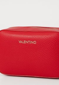 Raudona odinė kosmetinė su tekstūruotu paviršiumi, auksinėmis detalėmis ir įspaustu "VALENTINO" logotipu priekyje. Stačiakampio formos, užsegama užtrauktuku.