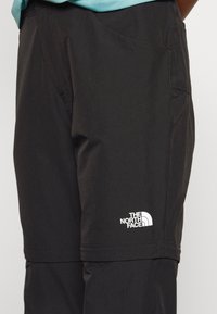 Zwarte outdoorbroek van duurzaam stof, met een zijzak, gestikte details en een wit logo op het onderbeen.