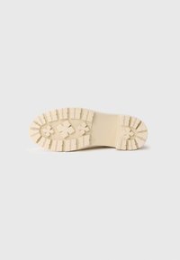 Semelle de chaussure en caoutchouc de couleur crème avec un motif de traction texturé, comportant des formes carrées et croisées pour l'accroche. Bord supérieur lisse.