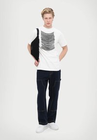 Balta kokvilnas t-kreklā ar melnu lineāru grafiku, apvienota ar tumši ziliem biksēm ar oranžiem diegiem un baltām kedām.