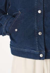 Giacca in denim blu scuro con bottoni a pressione argentati, due tasche laterali e dettagli di cucitura visibili. Tessuto testurizzato, design corto e aderente.