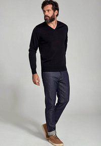Pull noir à col en V associé à un jean foncé, affichant une coupe ajustée. Le modèle porte des baskets beige avec les jambes du jean retroussées.