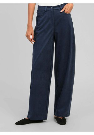 Femme portant un pantalon large bleu marine et des chaussures plates noires, debout avec une main dans la poche et l'autre détendue sur le côté.