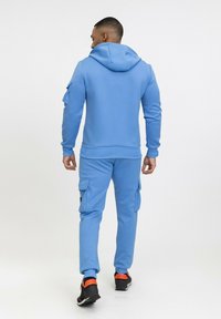 Sweat à capuche bleu avec cordon de serrage et pantalon cargo assorti, doté de poches et de poignets élastiques. Porté avec des baskets noires et oranges.