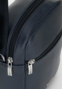 Borsa di un blu navy testurizzato con doppi zip, hardware liscio e forma arrotondata. Presenta un logo del marchio prominente su una piccola placca d'argento.