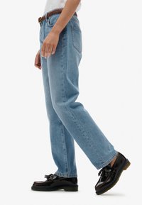 Jeans rectos de color azul claro con costuras visibles y cinco bolsillos, combinados con mocasines negros con detalle de lazo y un cinturón marrón texturizado.