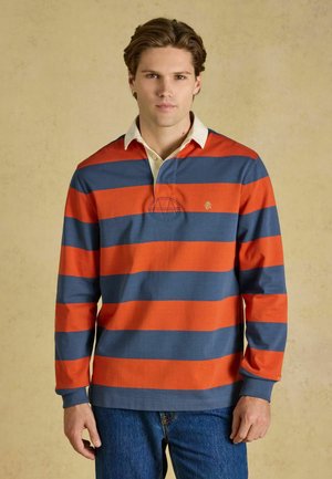 Giovane uomo con capelli castani corti indossa una maglia da rugby a maniche lunghe con larghe strisce orizzontali arancioni e blu e jeans blu.