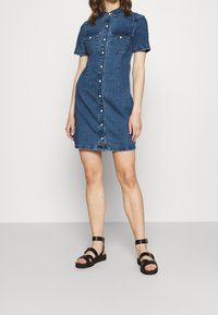 Denim skjortklänning i mörkblå, kort ärm, med knäppning framtill, två bröstfickor och en figursydd midja. Matchad med svarta sandaletter med remmar.