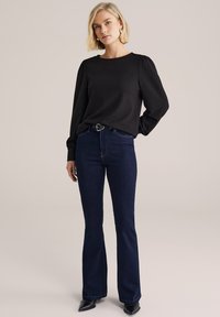 Rövid szőke hajú nő fekete hosszú ujjú felsőben, magas derekú sötétkék farmerral és fekete hegyes cipőben, sima háttér előtt állva.