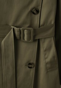 Gros plan d'un trench-coat vert olive avec boucle de ceinture rectangulaire recouverte de tissu et deux boutons foncés visibles à l'avant.