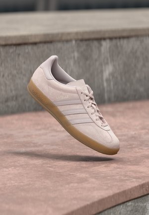 Gaiši rozā zamšādas sporta kurpes ar gumijas zoli. Tām ir trīs sānu svītras un iespiedums ar uzrakstu "Gazelle" sānu pusē.